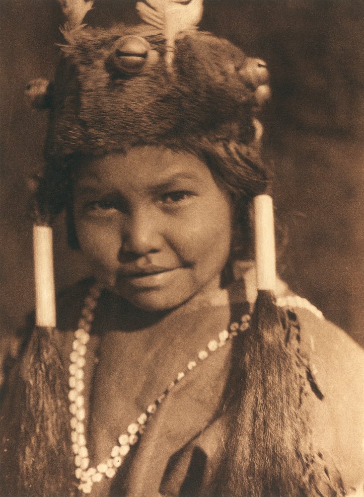 Close-up of an American Indian boy of the Klamath tribe or village. He is wearing a fur hat with applied acorns and feathers and wears his hair in two ponytails by Edward Sheriff Curtis
