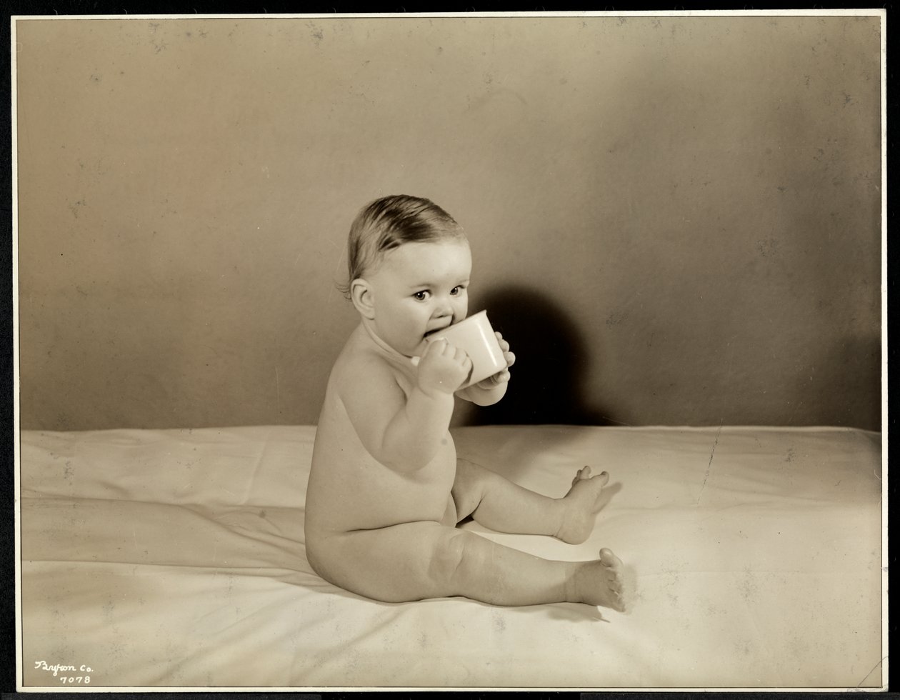 A Baby Sitting Up in Bed Drinking from a Cup at the Maternity Center Association at 70th Street East River, New York, 1935 by Byron Company