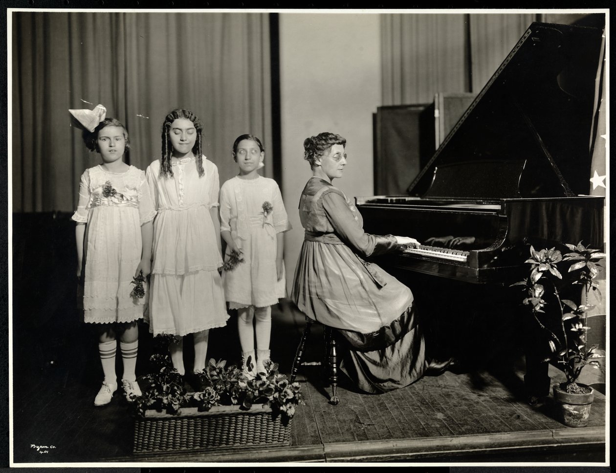 Woman playing the piano while three girls sing at the New York Association for the Blind, 111 East 59th Street, New York, c.1917 by Byron Company