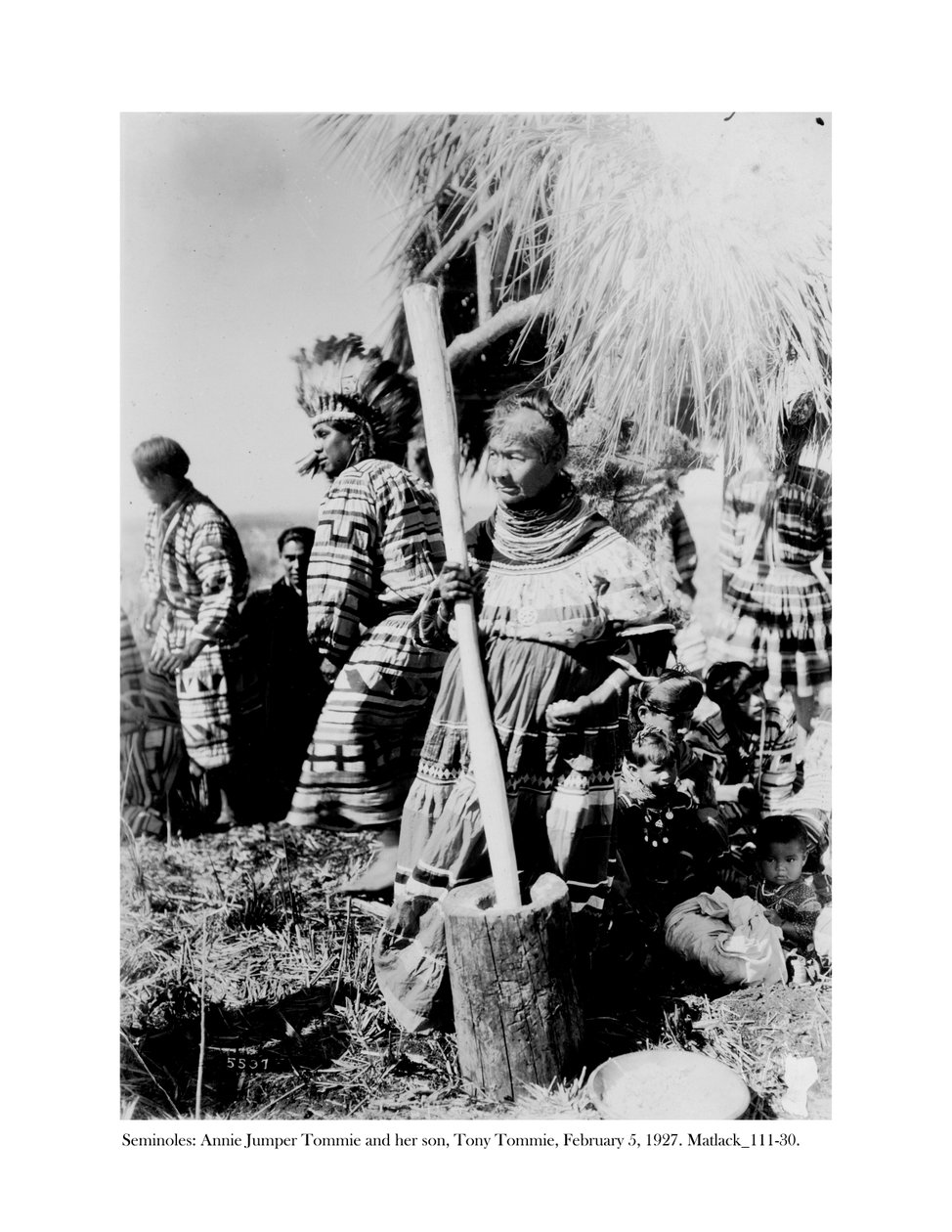 Annie Jumper Tommie Using a Mortar and Pestle to Make Cornmeal, and Her Son, Tony Tommie, Wearing an Eagle Feather Bonnet by Claude C. Matlack