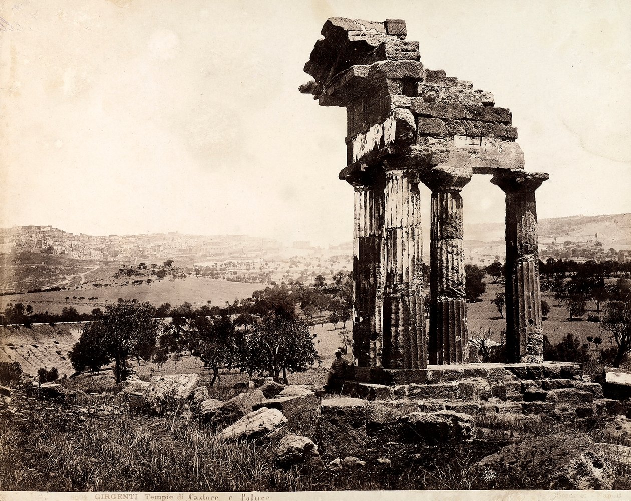 View of the remaining four columns supporting an entablature of the Temple of Castor and Pollux in the Valle dei Templi, Agrigento. A panorama of the city is visible in the background by Giorgio Sommer