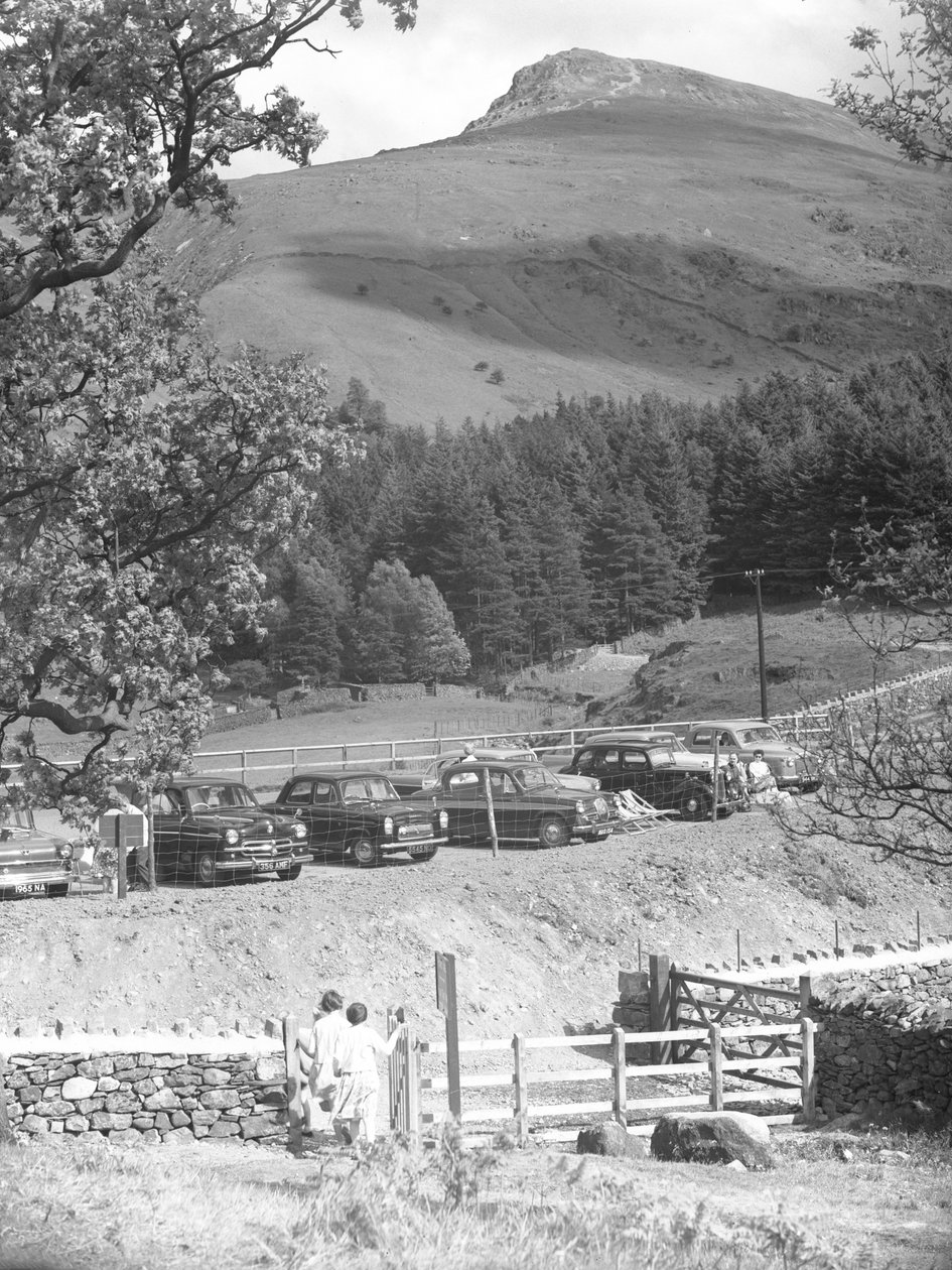 Two girls walking through a gate towards parked cars with hill behind by Joseph Hardman