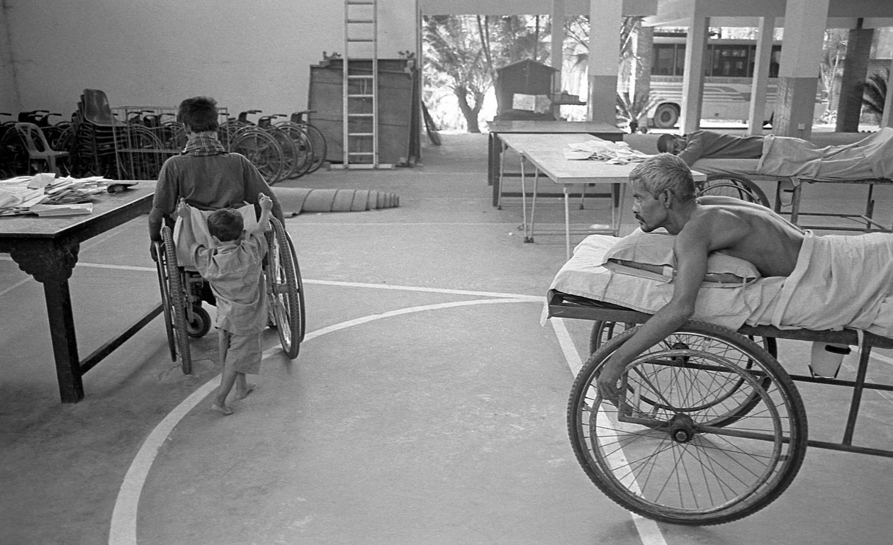 A physically challenged man on a wheelchair is carried by his son; another man with the same plight is watching at the Centre for the Rehabilitation of the Paralysed, CRP at Savar, Dhaka, Bangladesh, 2005 by Unbekannt Unbekannt