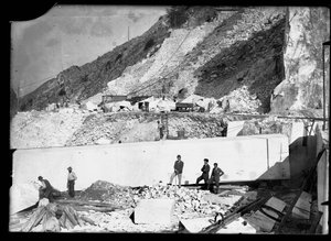 Three men observe the work of several excavators in the Apuan Alps. Behind them is an enormous block of marble already squared off and ready to be transported to the valley