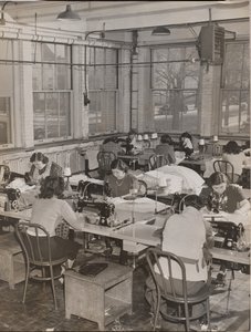 Women Seated at Desks Work on Sewing Machines