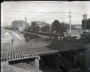 Horizontal, Black and White Photograph Showing a Wooden Bridge and Trolley Tracks over Partially Constructed Railroad Tracks. A Streetcar Marked Chouteau 2137 is Starting to Cross the Bridge on the Far Right. In the Distance Factory Buildings Reading Star by Charles Clement Holt