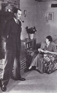 Wife reads, while husband warms his back against the fire by English Photographer