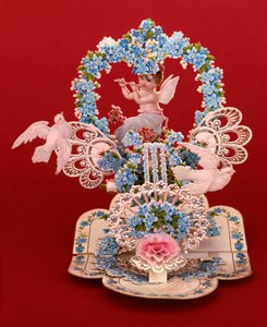 A pop-up card a winged cherub playing a flute under a heart shaped arch of forget-me-nots