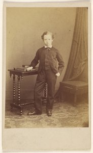 Unidentified little boy in suit standing at a table with books on top by Joseph Brown