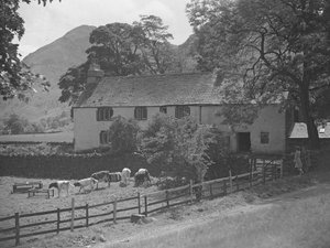 A building in front of which there are calves grazing, to the right two women stood talking against a wall and hills in the background