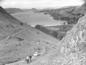 A view looking down a fellside with road, cars and people on, lake in background