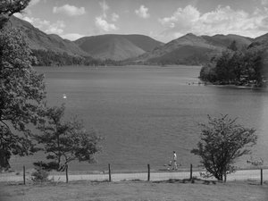 A lady looking across a lake, cars parked with people playing in the water, hills in background