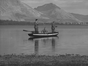 Three men in a rowing boat on a lake fishing, one man is seated, hills in background