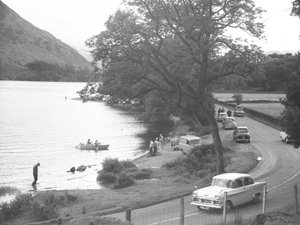 View of lake shore with people standing, walking, wading and boating, road runs to right of image with parked cars
