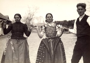 Two ladies and a man, in Hungarian traditional dresses, are holding hands in a dance, holding some candles in their hands by Rudolf Balogh