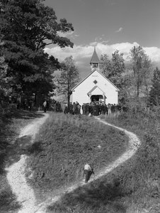 Men gathered outside of a small rural building, a schoolhouse or a church with bell