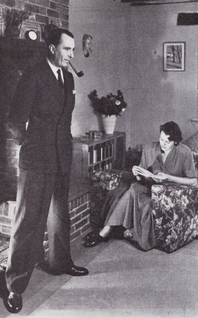 Wife reads, while husband warms his back against the fire by English Photographer