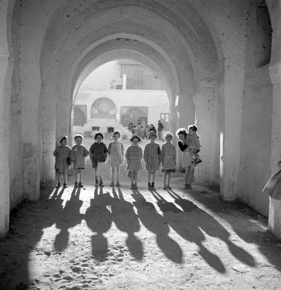 European children in school uniform under arch with shadows by Maynard Owen Williams