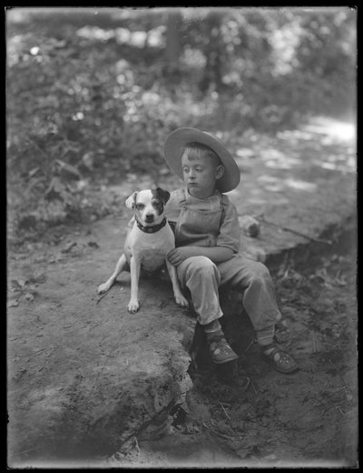 William Gray Hassler in straw hat and overalls, posed seated in the woods with a small dog (Bessie) by William Davis Hassler