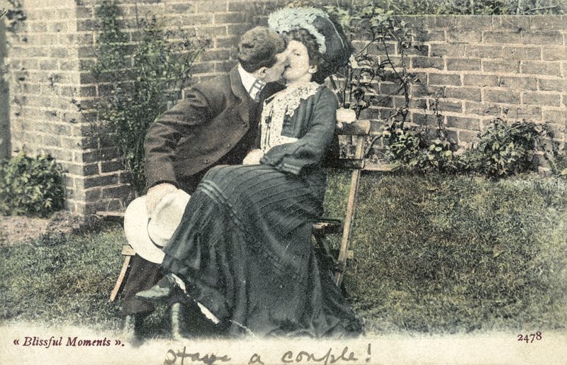 Man grabbing a kiss from a girl on a garden bench by English Photographer