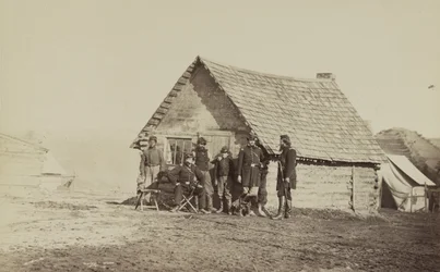 A group of soldiers, and two young men, one an African American, stand outside of log cabin quarters