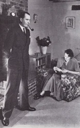 Wife reads, while husband warms his back against the fire