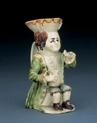 Toper Toby jug, his hat with brown and yellow scalloped edge, his incised brown hair en queue and with a white rosette, seated with a baluster jug on his knee, c. 1770