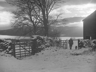 Man and dog walking through gate towards camera in snow with Thirlmere behind