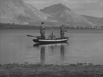 Three men in a rowing boat on a lake fishing, one man is seated, hills in background