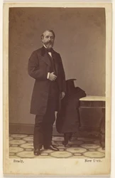 [Unidentified bearded man standing, a top hat and coat on nearby table]