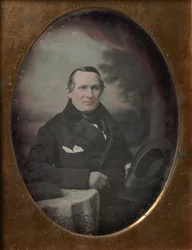 Man Wearing Coat and Gloves, Holding Hat, Seated in Front of Painted Outdoor Backdrop, 1840s