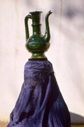 Woman in Kabul secluded from view by silk chadri carries jug