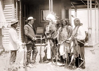 Three Cheyenne dressed in ceremonial costume and wearing guns greet a White accompanied by an interpreter