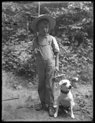 William Gray Hassler in straw hat and overalls, carrying fishing pole and basket, posed in the woods with a small dog (Bessie)